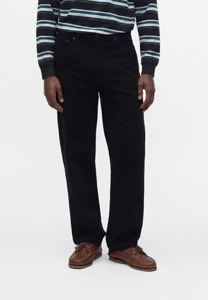 Homme portant un pantalon noir droit, des chaussures bateau en cuir marron et une chemise rayée sombre à manches longues, debout devant un fond blanc.