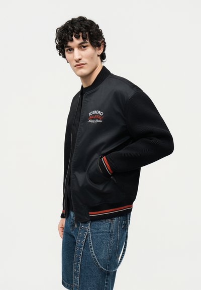 Νεαρός άνδρας με σγουρά μαλλιά, φοράει μαύρο μπουφάν τύπου bomber με το λογότυπο «Iceberg» και μπλε τζιν σορτς, σε απλό φόντο.