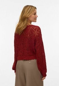 Pullover rosso lavorato a maglia con un motivo a pizzo, maniche lunghe e orlo scallopato. Indossato sopra pantaloni beige, che mettono in evidenza i dettagli traforati.