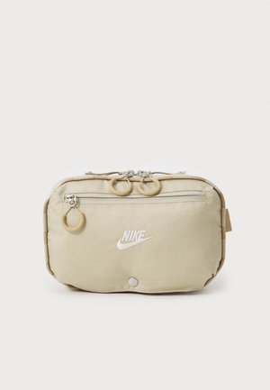 Beige Nike heuptas met voorvak met ritssluiting, wit geborduurd logo en ronde ritstrekkers tegen een effen achtergrond.