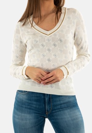 Kvinde iført en hvid V-hals sweater med sølvblomster og guldkant, kombineret med blå denimjeans, hænderne foldet ved livet.