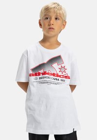 Blond chlapec stojí s rukami za chrbtom, oblečený v bielej tričku DC Shoes s veľkým čiernym a červeným grafickým textom vpredu.