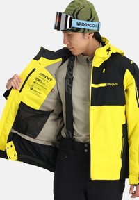 Wasserabweisende Spyder-Ski-Jacke in leuchtendem Gelb und Schwarz, ausgestattet mit einem Reißverschluss vorne, einer inneren Netz-Tasche und einer verstellbaren Kapuze.