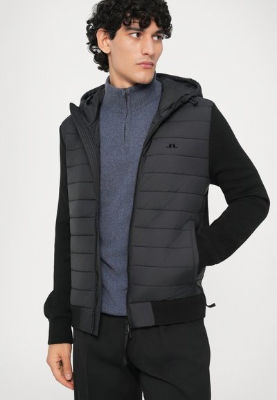 Casaco híbrido masculino com a parte frontal acolchoada preta, capuz e mangas de malha preta. Usado sobre um suéter azul com fecho zip, com um detalhe de logo discreto.