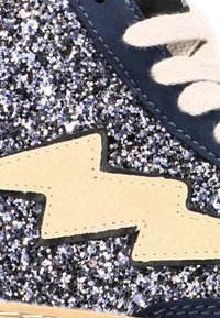 Zapatilla azul oscuro de lado con brillo plateado, parche de rayo beige y cordones en blanco roto visibles.