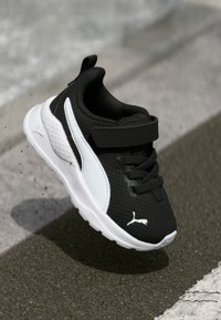 Baskets noires et blanches avec empeigne en mesh, col rembourré et sangle Velcro. Présente un logo prominent sur le côté et une semelle en caoutchouc.