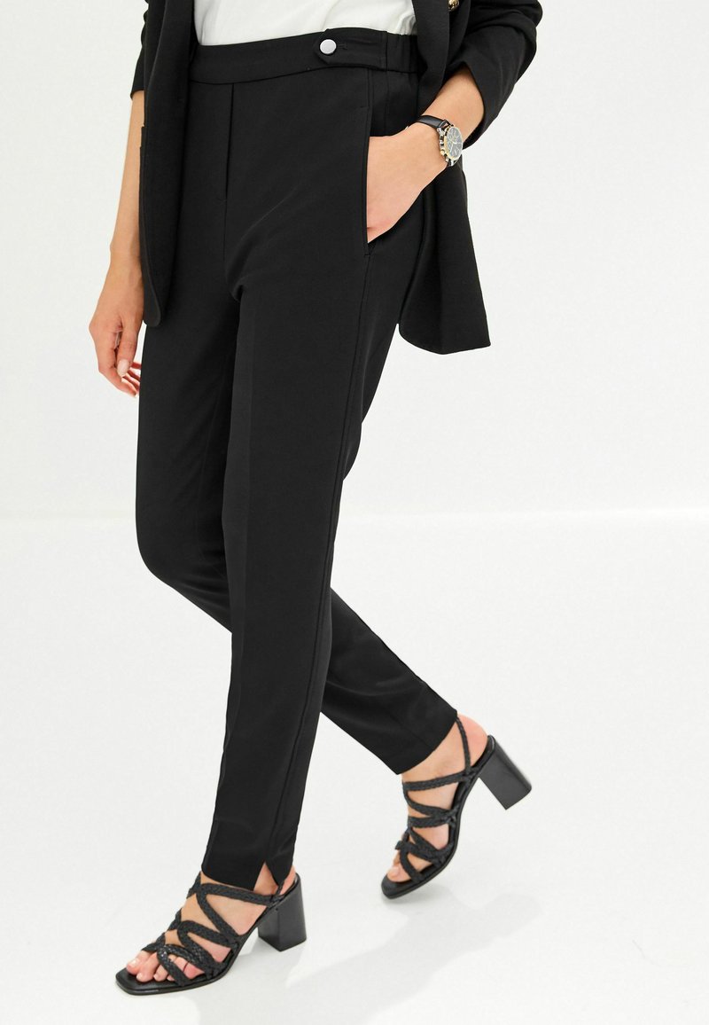 Pantalon noir ajusté avec une texture lisse, doté de poches latérales et d'un bouton à l'avant. Assorti à des sandales noires à talons carrés avec des lanières.