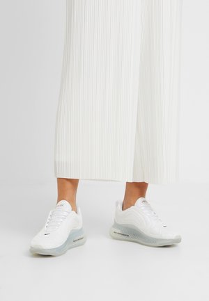Person iført hvide plissee wide-leg bukser og hvide atletiske sneakers med gennemsigtige såler og tekstdetaljer på siden.