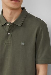 Olivgrünes Poloshirt aus strukturiertem Stoff mit Kentkragen, ausgestattet mit einem kleinen Logo-Patch auf der linken Brust und einer Knopfleiste.