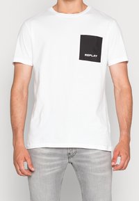 Bílý tričko s krátkým rukávem a černou čtvercovou kapsou. Na kapse je vytištěno logo "REPLAY". Hladká tkanina, standardní design s kulatým výstřihem.