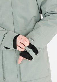 Ljusgrön, texturerad vinterjacka med justerbar manschettstängning och inre svart foder. Har en zippered ficka på den nedre sidan.