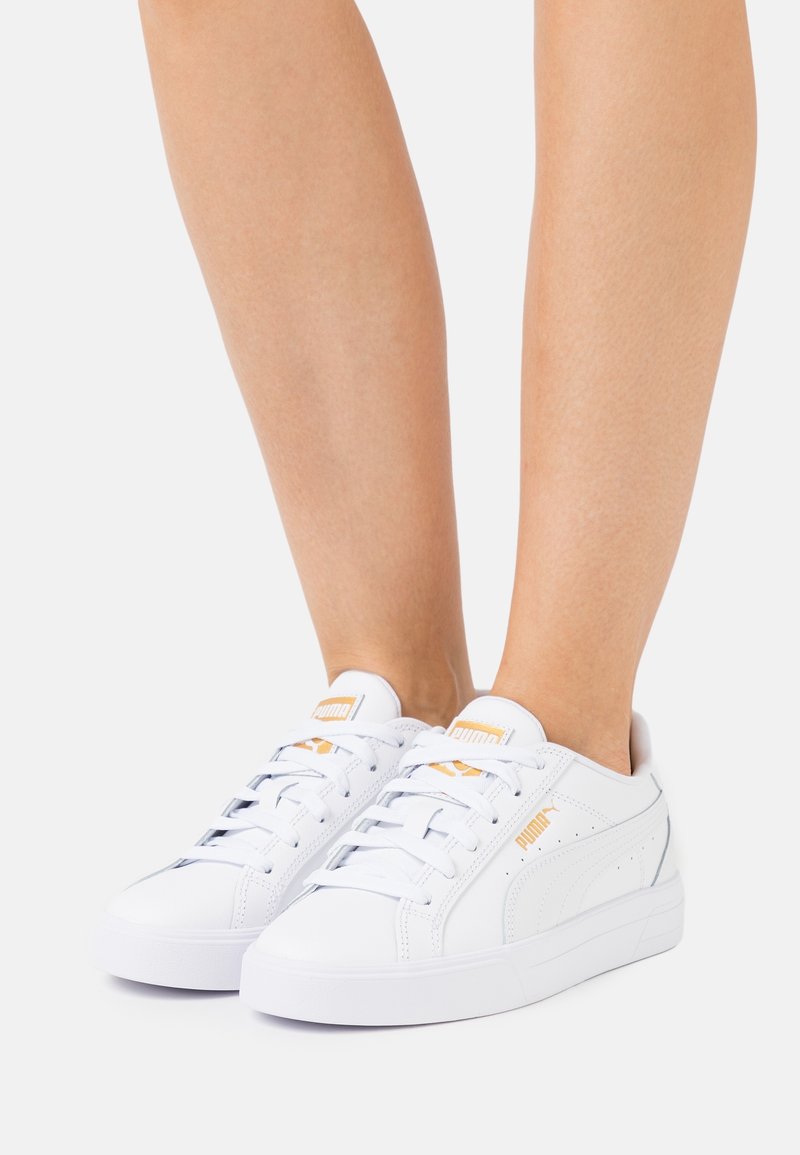 Personne portant des baskets blanches Puma avec des logos dorés, montrée de la moitié du mollet vers le bas sur un fond clair uni.