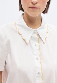 Femme portant un chemisier blanc à manches courtes avec des bords festonnés et des surpiqûres jaunes le long du col et de la patte de boutonnage.