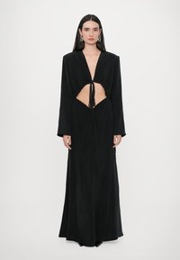 Robe longue noire avec un décolleté en V profond, section médiane ouverte avec un lien, et manches longues. Tissu lisse et fluide avec un design minimaliste.