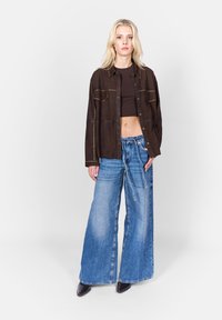 Brun mocka skjortjacka med två bröstfickor och metallnitar, kombinerad med vida blå denimbyxor och mörka skor.