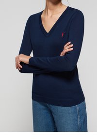 Pull-over en tricot bleu marine avec un col en V, des manches longues et un ourlet côtelé. Il présente un petit logo rouge sur la poitrine gauche. Texture douce.