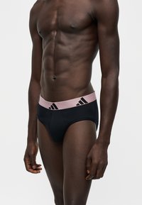 Мускулест мъж, носещ черни слип с розова талия с черно лого на Adidas, стоящ на светъл фон.