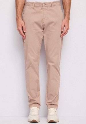 Uomo con chinos beige e sneakers bianche in piedi davanti a uno sfondo chiaro uniforme, con le mani rilassate lungo i fianchi.