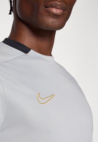 Γκρι αθλητική μπλούζα με μαύρο γιακά, που διαθέτει χρυσό κεντητό λογότυπο στο στήθος. Λείο υφή, σχεδιασμένη για αθλητική χρήση.