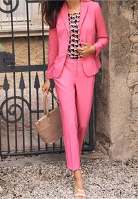 Rosa skräddarsydd kavaj och byxor med en mönstrad topp. Tillbehören inkluderar en beige toalettväska. Remsandaler fulländar outfiten.