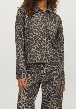 Femme portant une veste et un pantalon assortis avec un imprimé léopard marron et noir, main détendue le long du corps, cheveux bouclés visibles.