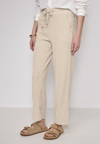 Beige vida byxor med texturerad yta, med dragsko i midjan och sidofickor, tillsammans med bruna slip-on sandaler.