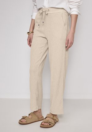 Beige weit geschnittene Hose mit strukturiertem Finish, ausgestattet mit einem Kordelzugbund und Seitentaschen, kombiniert mit braunen Slipper-Sandalen.