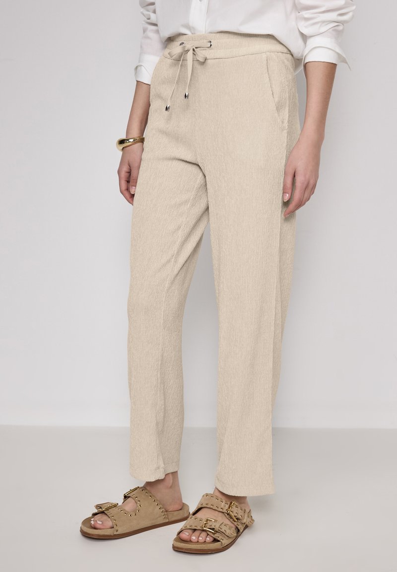 Beige vida byxor med texturerad yta, med dragsko i midjan och sidofickor, tillsammans med bruna slip-on sandaler.