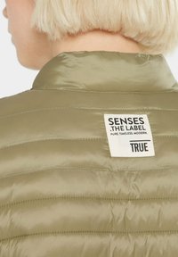 Olivgrüne Pufferjacke mit einer gesteppten Textur. Verfügt über einen Kragen und ein Etikett auf der Rückseite mit der Marke "SENSES THE LABEL".