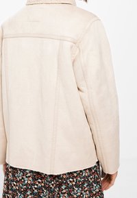 Beige suède jas met een schapenvachtkraag, rechte pasvorm en strakke lijnen. Voorzien van achternaden en een zachte textuur. Daaronder een rok met bloemmotief.