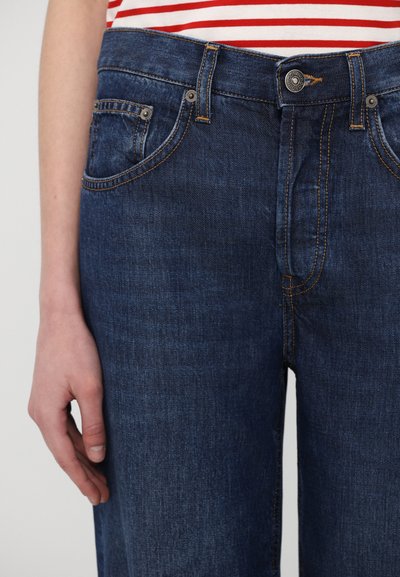 Tumedat sinised denim püksid, millel on nähtavad õmblused, nupuga sulgemine ja viie tasku disain. Kanga pind tundub sile, kuid kergelt tekstureeritud.
