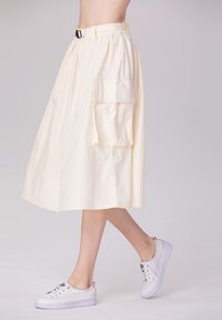 Jupe jaune clair, jusqu'aux genoux, en tissu doux, dotée d'une ceinture et d'une grande poche latérale. Associée à des baskets blanches.