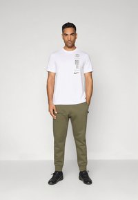 Balta īsroku T-krekls ar melniem grafiskiem elementiem, kopā ar olīvu zaļiem sporta biksēm. Abiem apģērba gabaliem ir Nike logo. Melni sporta apavi.