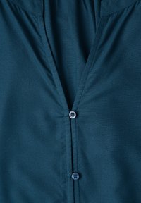 Devant de chemise en tissu vert sarcelle foncé avec deux boutons visibles, un fermé près du haut et un en dessous, montrant de légers plis autour de la zone des boutons.