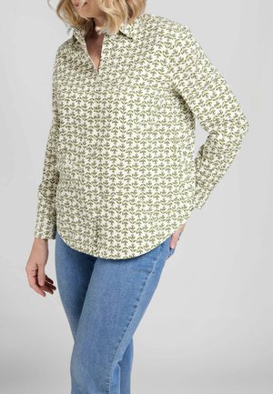 Donna che indossa una camicia bianca con bottoni e motivo a foglie verdi e jeans blu, in piedi con una mano in tasca contro uno sfondo grigio.