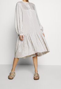 Lehké pruhované šaty s dlouhými rukávy, knoflíkovým detailem u výstřihu a sukýnkovým designem s volány. Barvy zahrnují světle modrou a bílou.