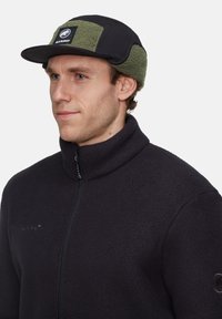 Sort fleecejakke med høj krave, som har en frontlynlås og logoet "MAMMUT." Bæres med en sort kasket med grønne fleecepaneler.