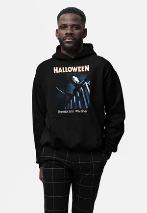 Mężczyzna ubrany w czarną bluzę z kapturem z napisem "HALLOWEEN" i obrazem osoby trzymającej nóż na schodach, w połączeniu z czarnymi spodnimi w kratę.