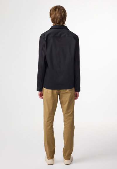 Svart långärmad skjorta med klassisk krage, ihop med ljusbruna chinos och beige skor, sett från baksidan mot en neutral bakgrund.