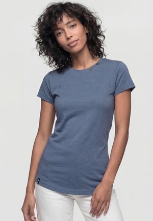 Frau mit lockigem dunklem Haar, die ein blaues kurzärmeliges T-Shirt und weiße Hose trägt, steht vor einem hellen einfarbigen Hintergrund.