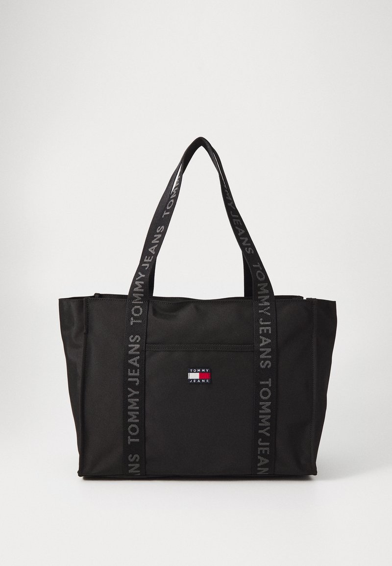 Fekete vászontáska tartós anyagból, elülső zsebbel, párhuzamos fogantyúkkal és a "TOMMY JEANS" ismétlődő mintájával díszített pántokkal.