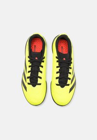 Botas de fútbol amarillas brillantes con acentos negros, que cuentan con una parte superior texturizada, cordones negros y un forro interior rojo.
