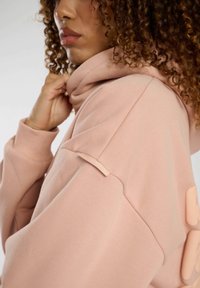 Ljusrosa hoodtröja med hög krage, texturerad tyg och sidetikett. Har en avslappnad passform och en subtil, mjuk silhuett.