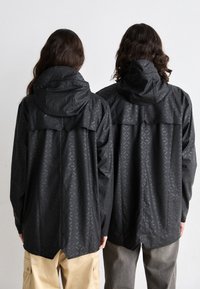 Des vestes noires imperméables avec capuche, arborant un motif géométrique subtil et un dos fendu, associées à un pantalon cargo beige clair.
