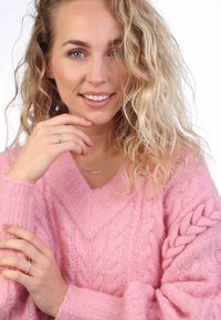 Rosa Kabelstrickpullover mit V-Ausschnitt, der geflochtene Akzente an den Ärmeln aufweist. Das Model trägt eine silberne Halskette und einen Ring, die eine strukturierte Optik präsentieren.