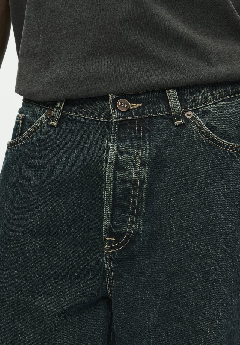 Dark green denim jeans with a visible button closure and stitching. Features side pockets and a classic fit design. Cotton material.