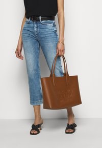 Braune Leder-Tote mit geprägtem Markenlogo, strukturiertem Design, doppelten Griffen und ohne sichtbare Beschläge, kombiniert mit blauen Jeans und schwarzen Sandalen.
