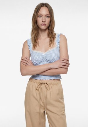 Giovane donna con capelli castano chiaro mossi, indossa un crop top a quadretti blu con bordatura in pizzo e pantaloni beige con coulisse, braccia incrociate.