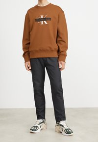 Sudadera marrón con el logo "Calvin Klein Jeans" en blanco, combinada con pantalones negros y zapatillas deportivas multicolores. Textura suave, ajuste casual.