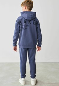 Námornícky modré mikina s kapucňou a zodpovedajúce nohavice, s výrazným potlačou "BAKER" na chrbte, jemná textúra a rebrované manžety. Biele tenisky so značkovaním.